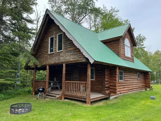 A log cabin at Papin's Resort with a green metal roof and a covered porch, a fire ring in the grass out front and pines behind.
